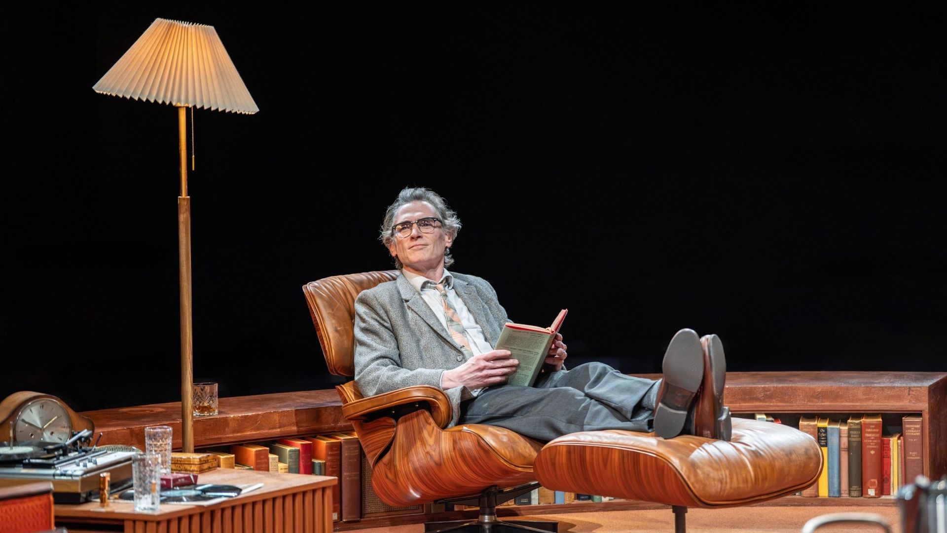 Man in grey suit reading book, reclining on brown leather chair, stage setting, dark background.