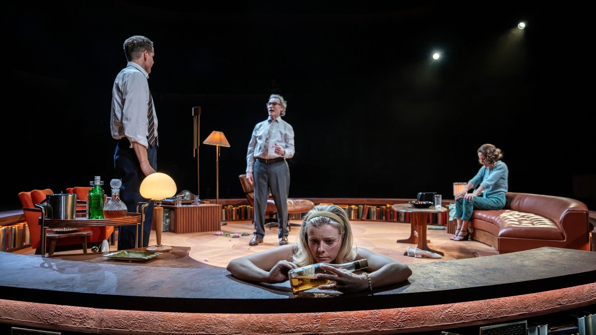 Three actors perform on stage; one woman lies down, two men stand, surrounded by vintage furniture.