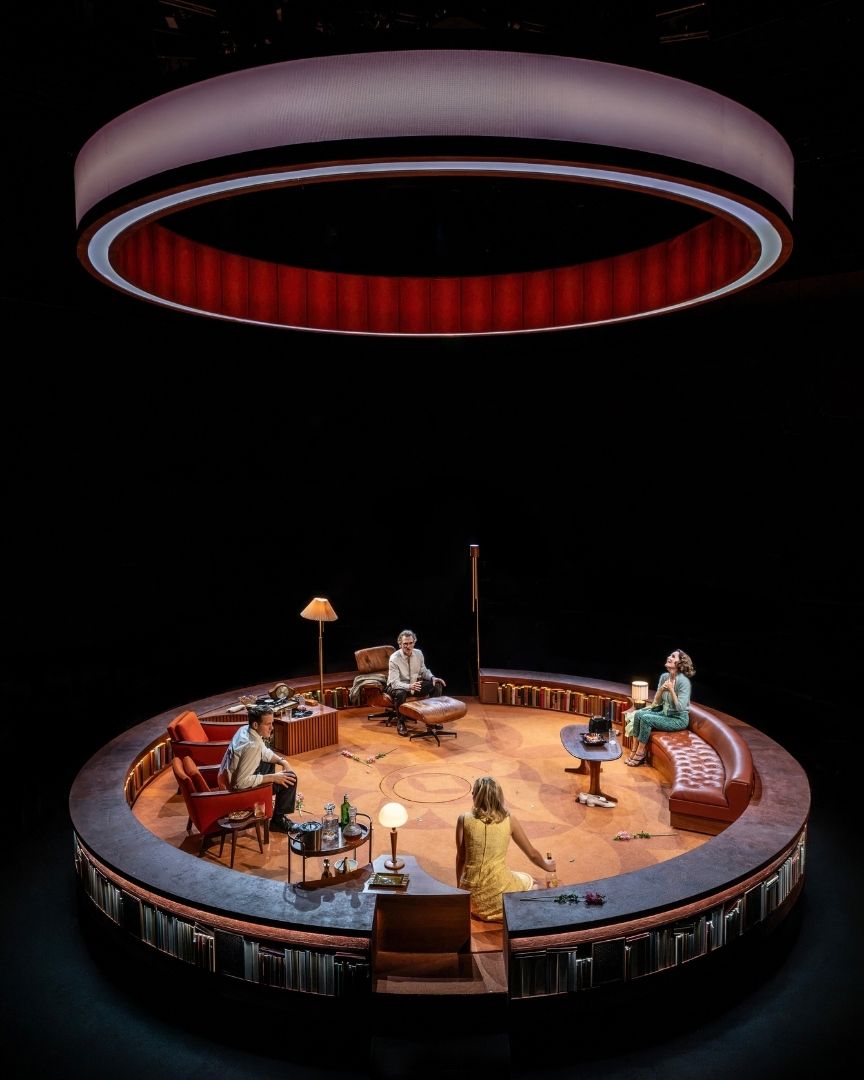 Four actors on a circular stage set with vintage furniture, under a large circular light.