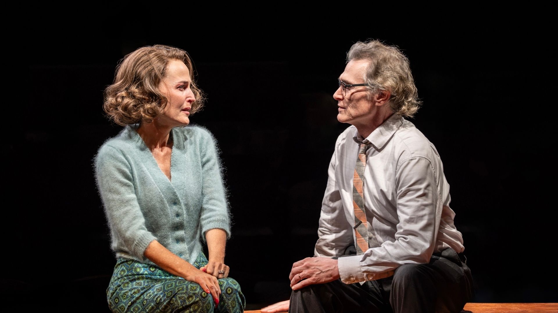 Two actors in conversation on stage; woman in turquoise cardigan, man in white shirt and tie.