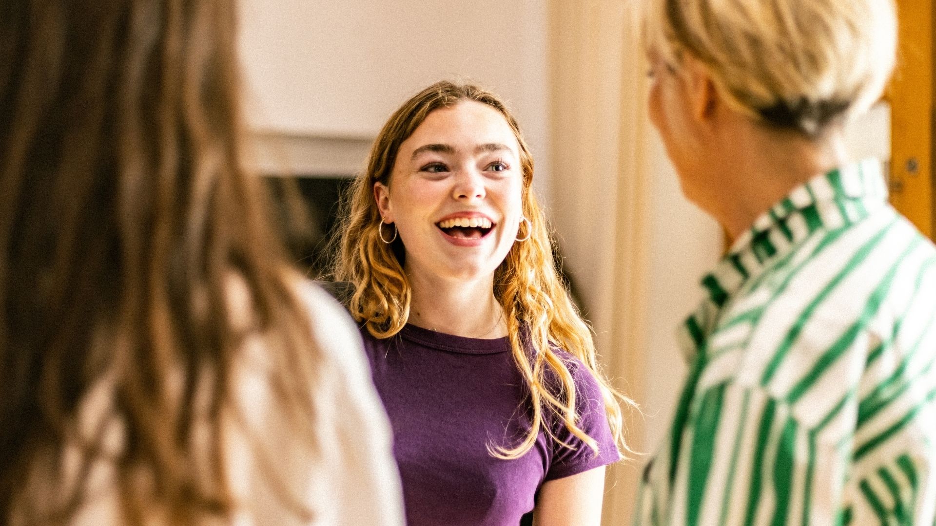 A young woman in a purple shirt smiles while talking to two others indoors.