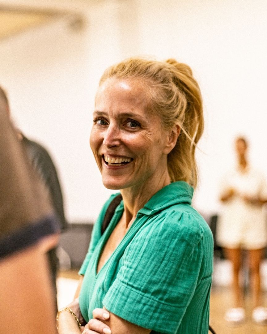 Smiling woman with blonde hair in a green shirt, standing indoors with a blurred background.