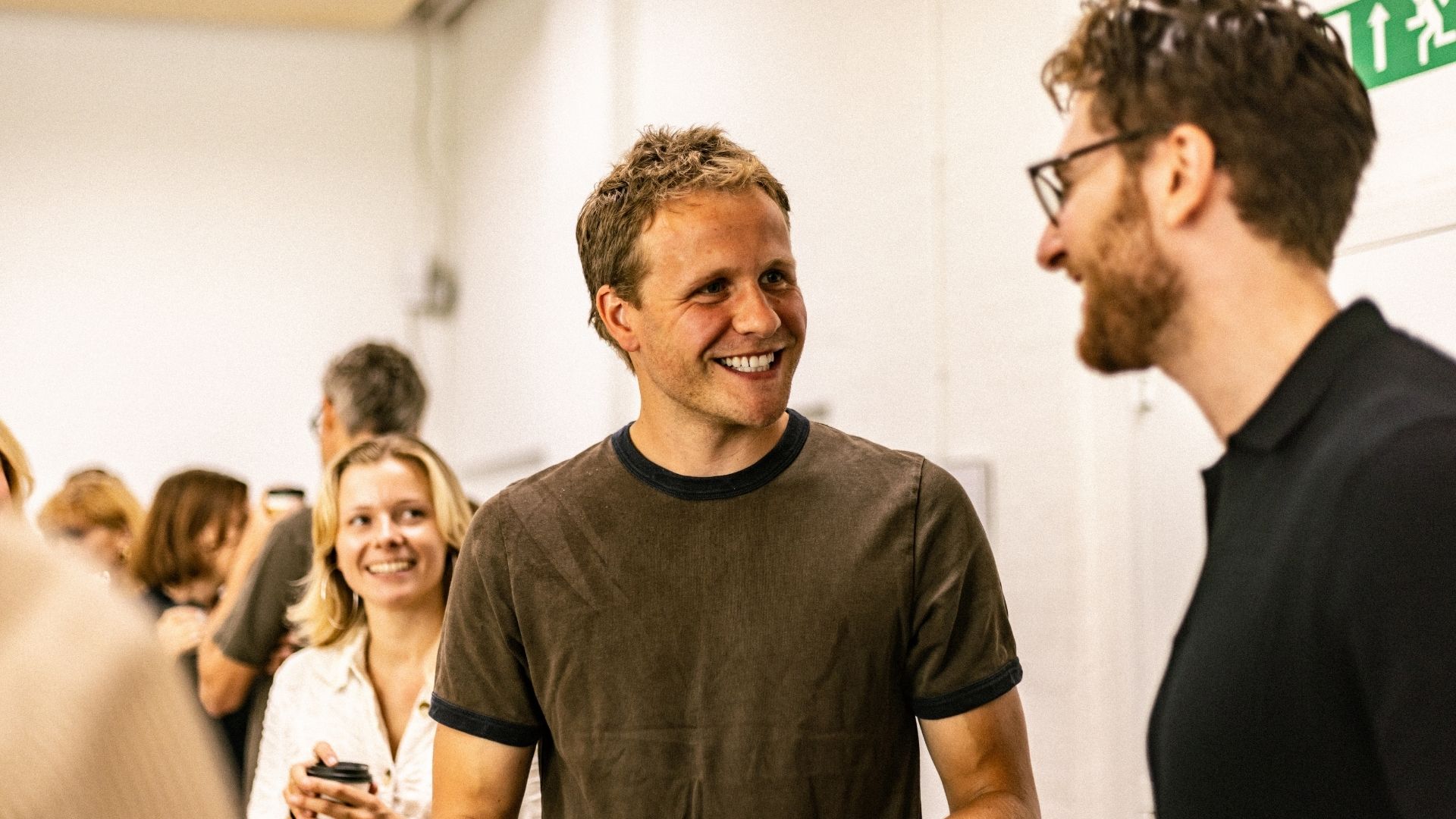 A man in a dark t-shirt smiles while talking to another man.