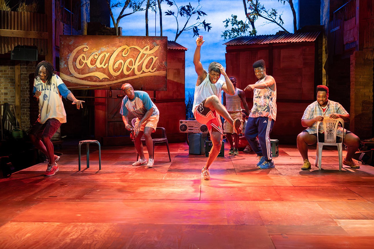 Performers energetically dance on stage with vibrant lighting; a Coca-Cola sign is visible in background.