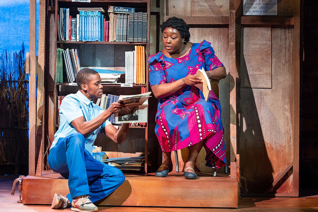 A man in blue and a woman in a vibrant dress sit by a bookshelf, reading.