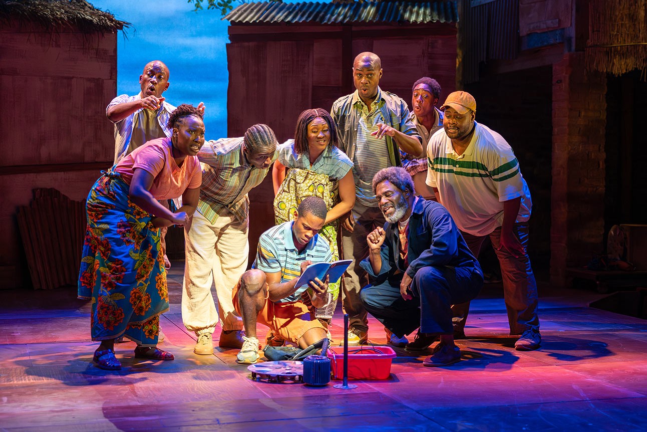 A group of actors on stage, gathered around a boy reading a book, colourful costumes.