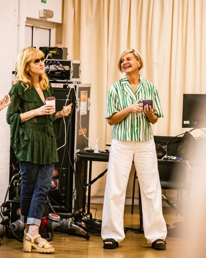 Two women smiling, holding mugs, in a rehearsal room with electronic equipment and beige curtains.