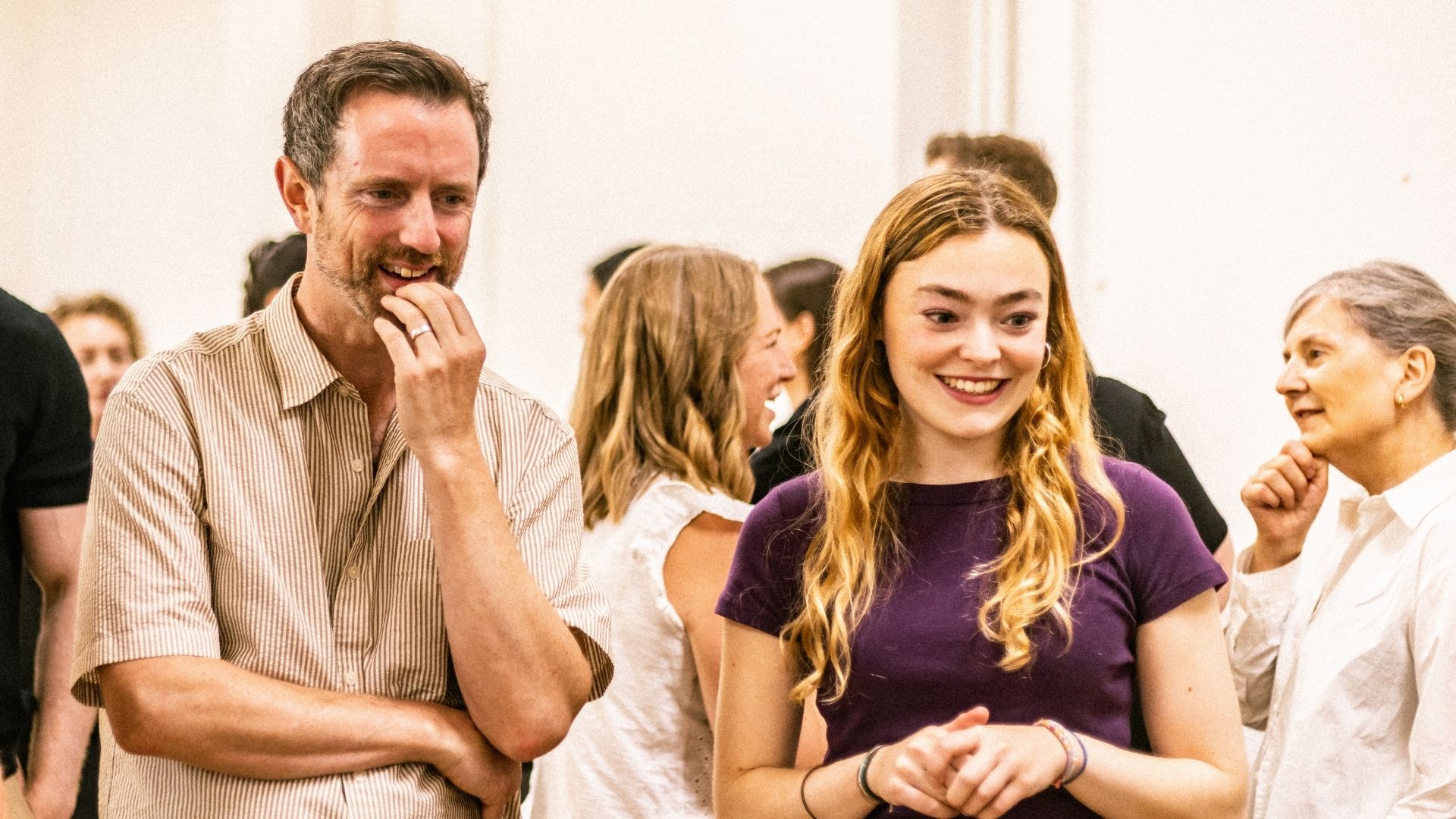 A man and woman smiling in conversation, surrounded by people in a brightly lit room.