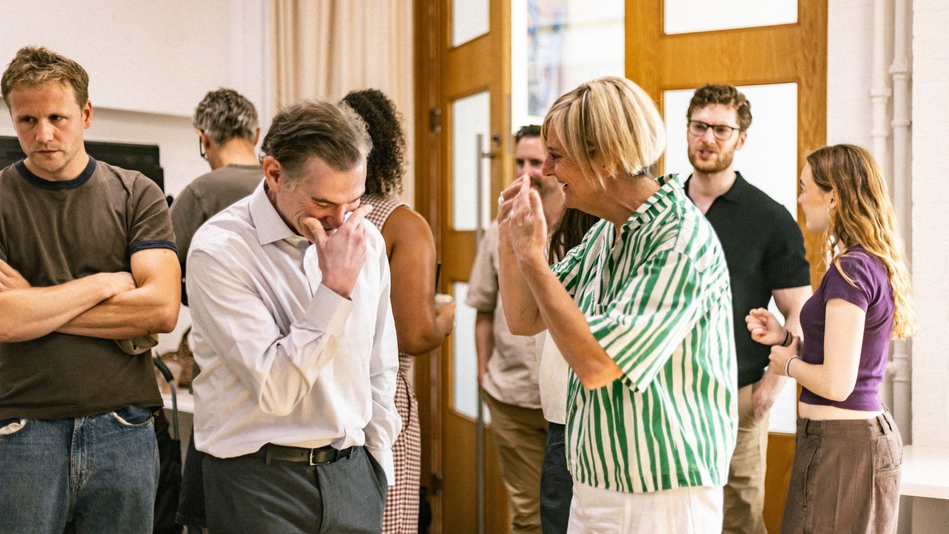 A group of people in conversation, one in a striped shirt, indoors with wooden doors visible.