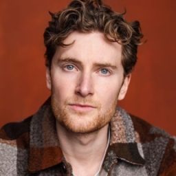 A man with curly hair and a plaid jacket poses against an orange background.
