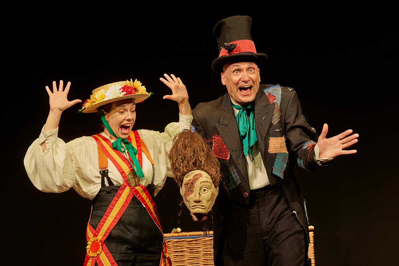 Two actors in colourful costumes perform energetically on stage, with a wicker basket and mask.
