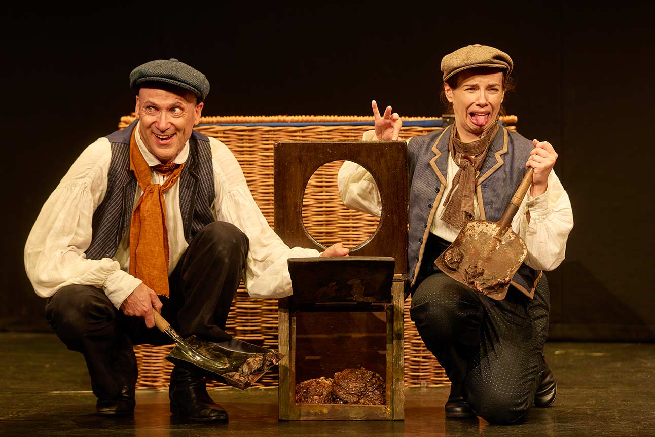 Two actors in period costumes pose humorously with a wooden toilet prop on stage.