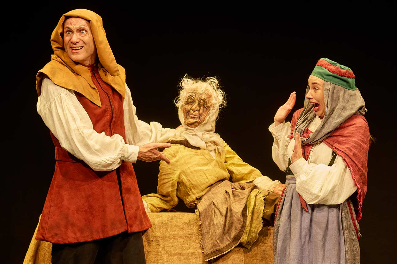 Two actors in medieval costumes perform energetically on stage, with a puppet in the centre.