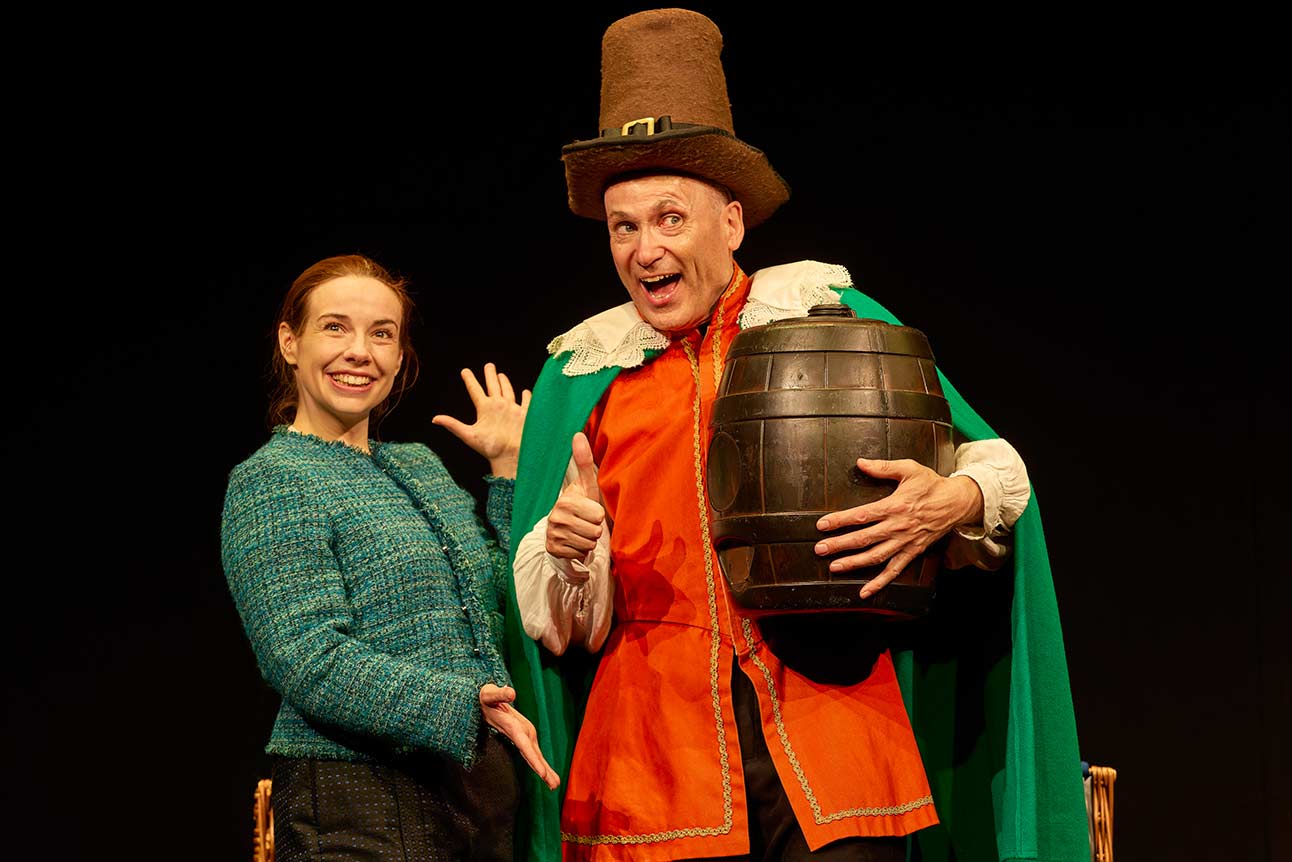 Two actors on stage; one in a green cloak and tall hat holding a barrel, smiling.