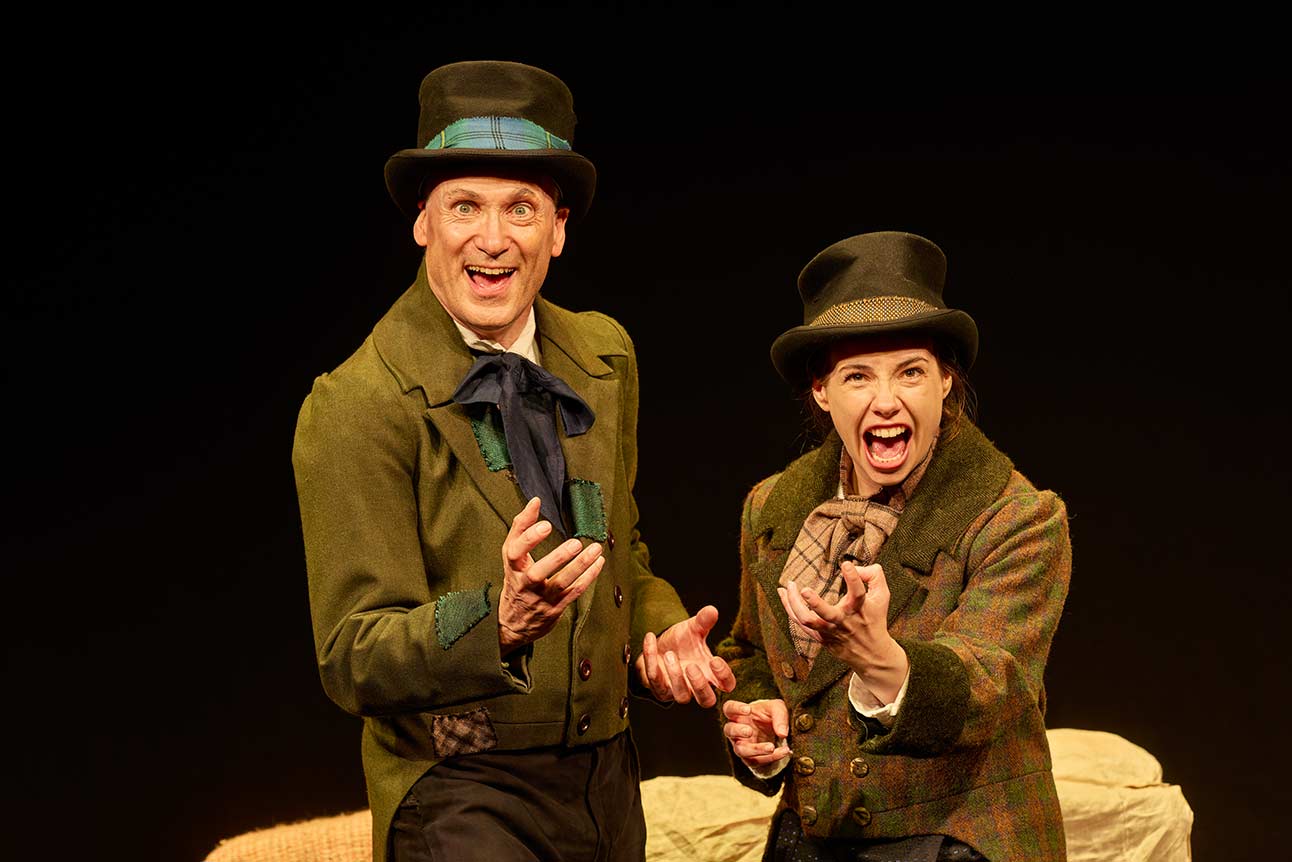 Two actors in Victorian costumes enthusiastically gesturing on stage, wearing green jackets and top hats.