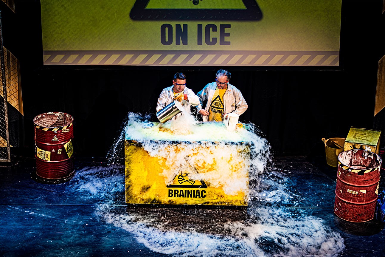 Two performers in lab coats on stage with colourful smoke and scientific equipment in the background.