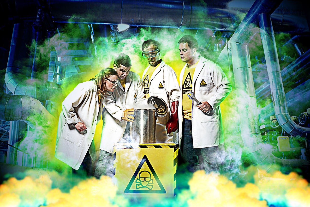 Four performers in lab coats conducting a experiment on stage with colourful green smoke effects.