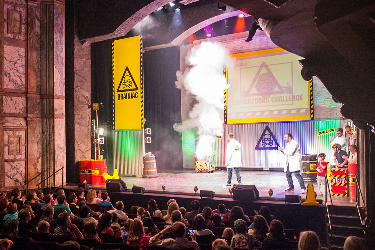Three performers in lab coats conducting a science experiment on stage with colourful smoke effects.