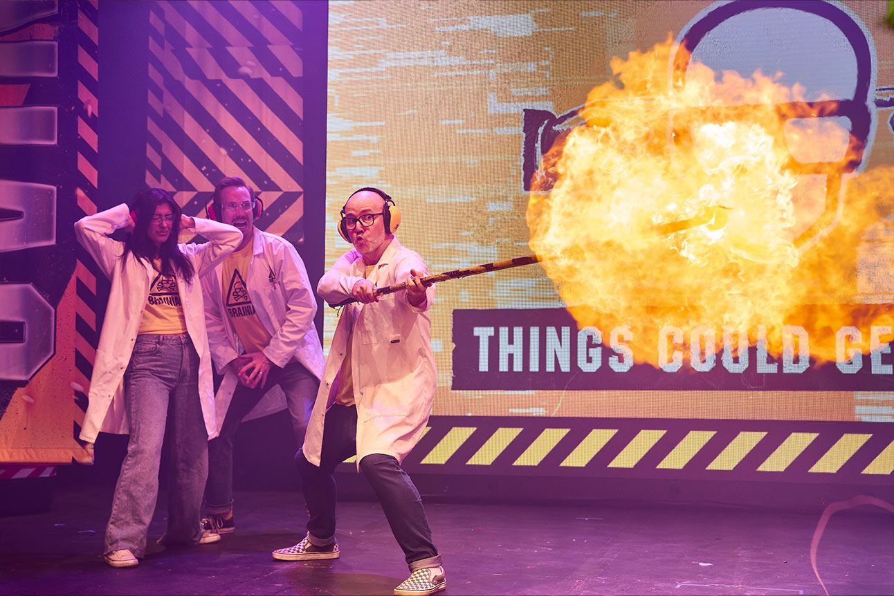 Two performers in lab coats, one holding a clipboard, stand on stage with colourful props.