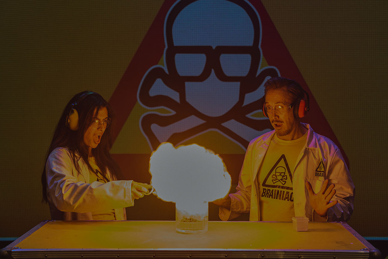 Two performers in lab coats conducting a colourful science experiment on stage with fire.