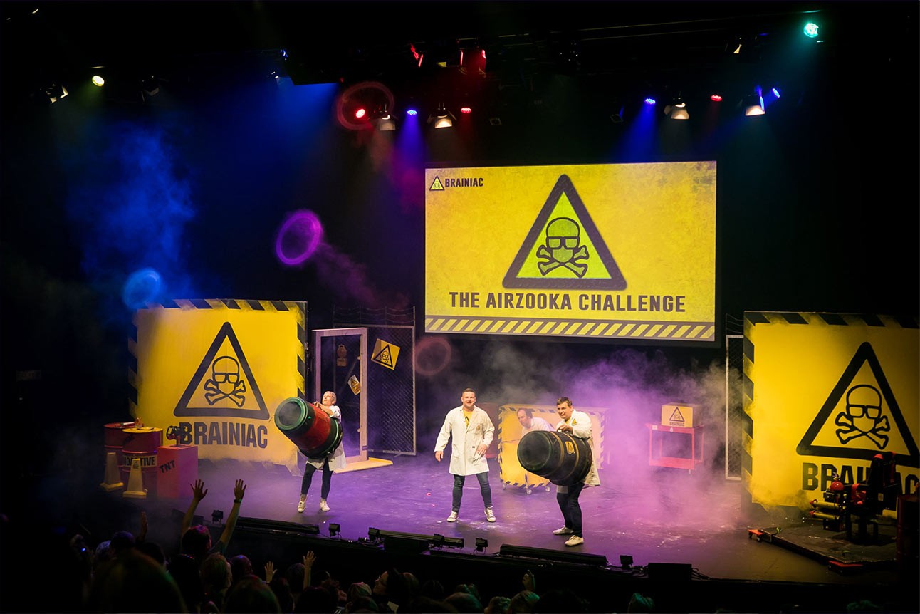 Three performers in lab coats energetically demonstrate a science experiment on stage with colourful props.