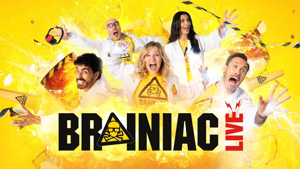 Five performers in lab coats on stage, surrounded by colourful fire and smoke, with a Brainiac Live banner.