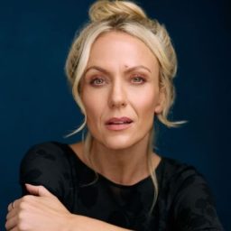 A woman with blonde hair in a bun poses against a blue background, wearing a black top.