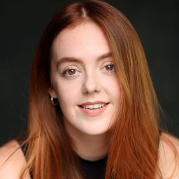 A woman with long red hair smiles against a dark background, wearing a black top.