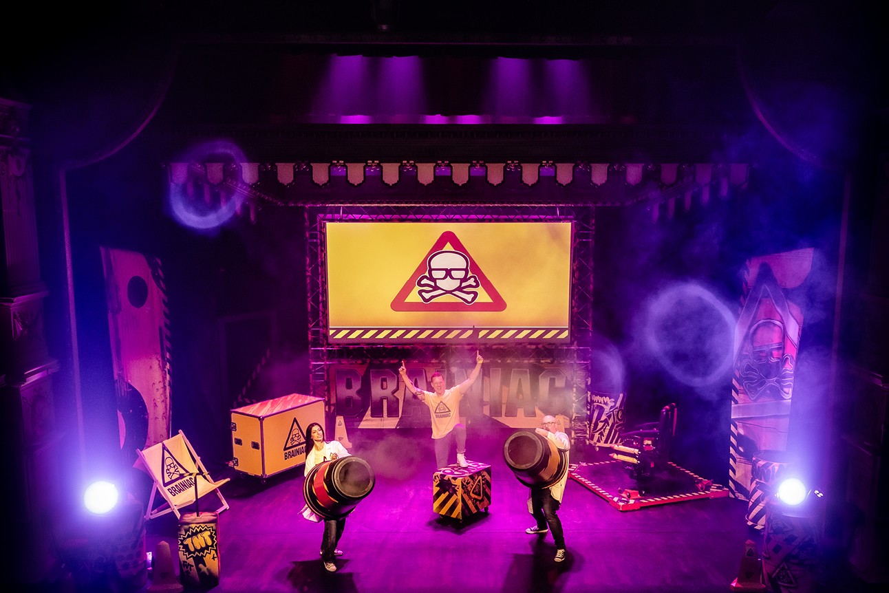 Three performers on stage with science-themed props, vibrant purple lighting, and a caution sign backdrop.