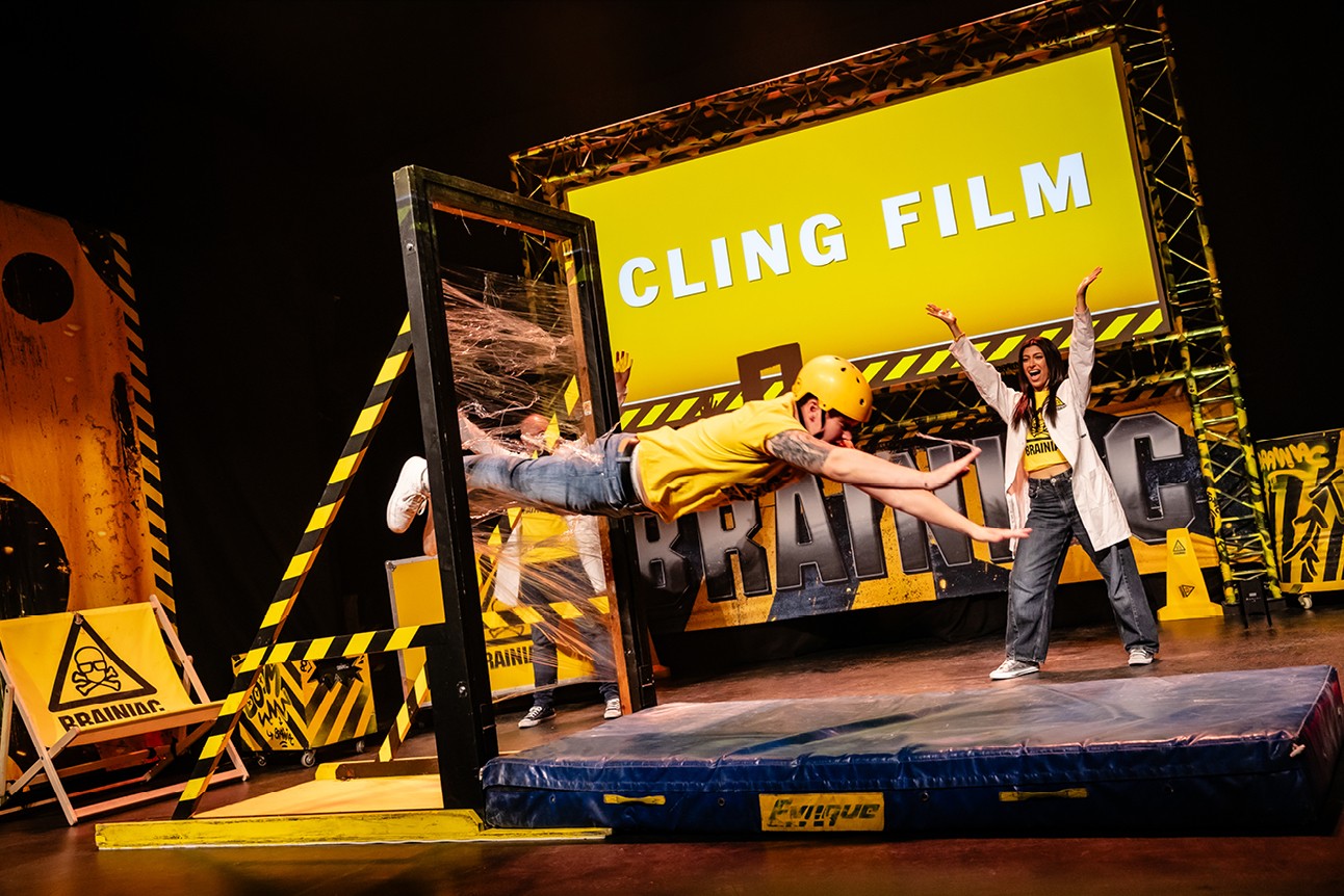 Performers in yellow outfits demonstrate a cling film stunt on stage, with a vibrant yellow backdrop.