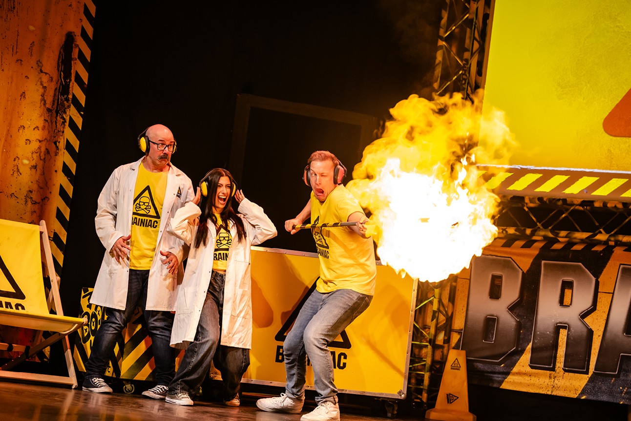 Three people in lab coats and yellow shirts react to a large fireball on stage.