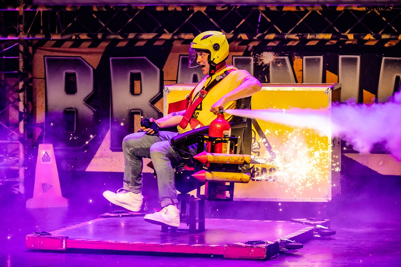 Person in yellow helmet and harness on spinning chair with sparks, purple stage lighting background.