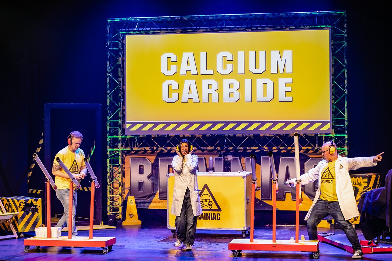 Three performers in lab coats on stage with "Calcium Carbide" sign, colourful props, and safety symbols.