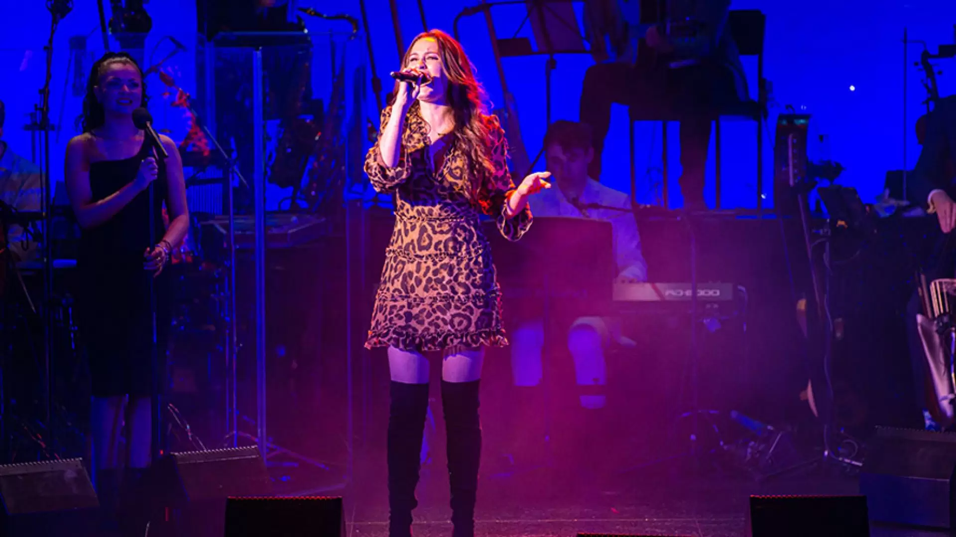 Singer in leopard print dress performs on stage, vibrant blue lighting, musicians in background.