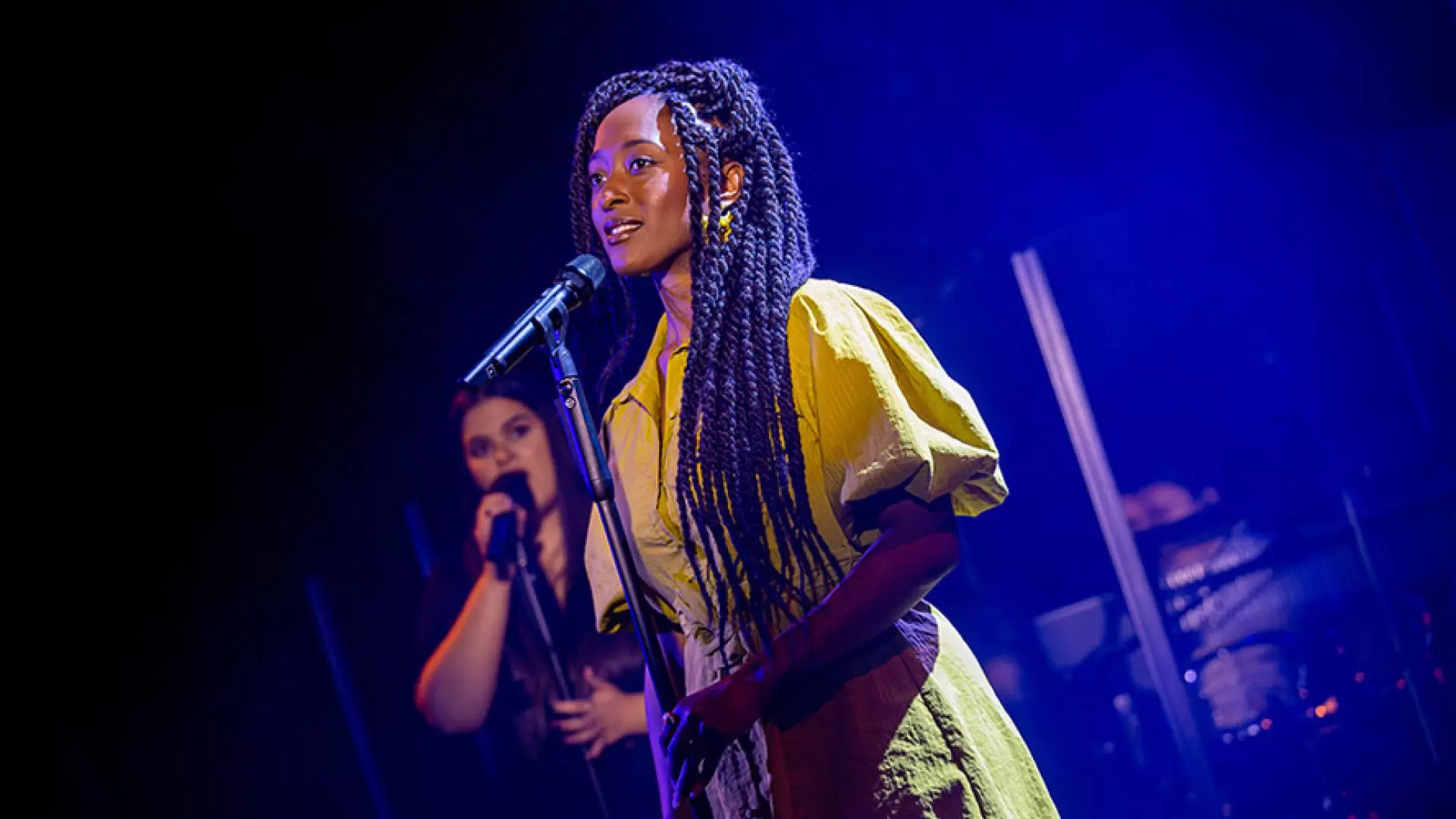 Two performers sing on stage; one in a yellow dress, the other in the background.