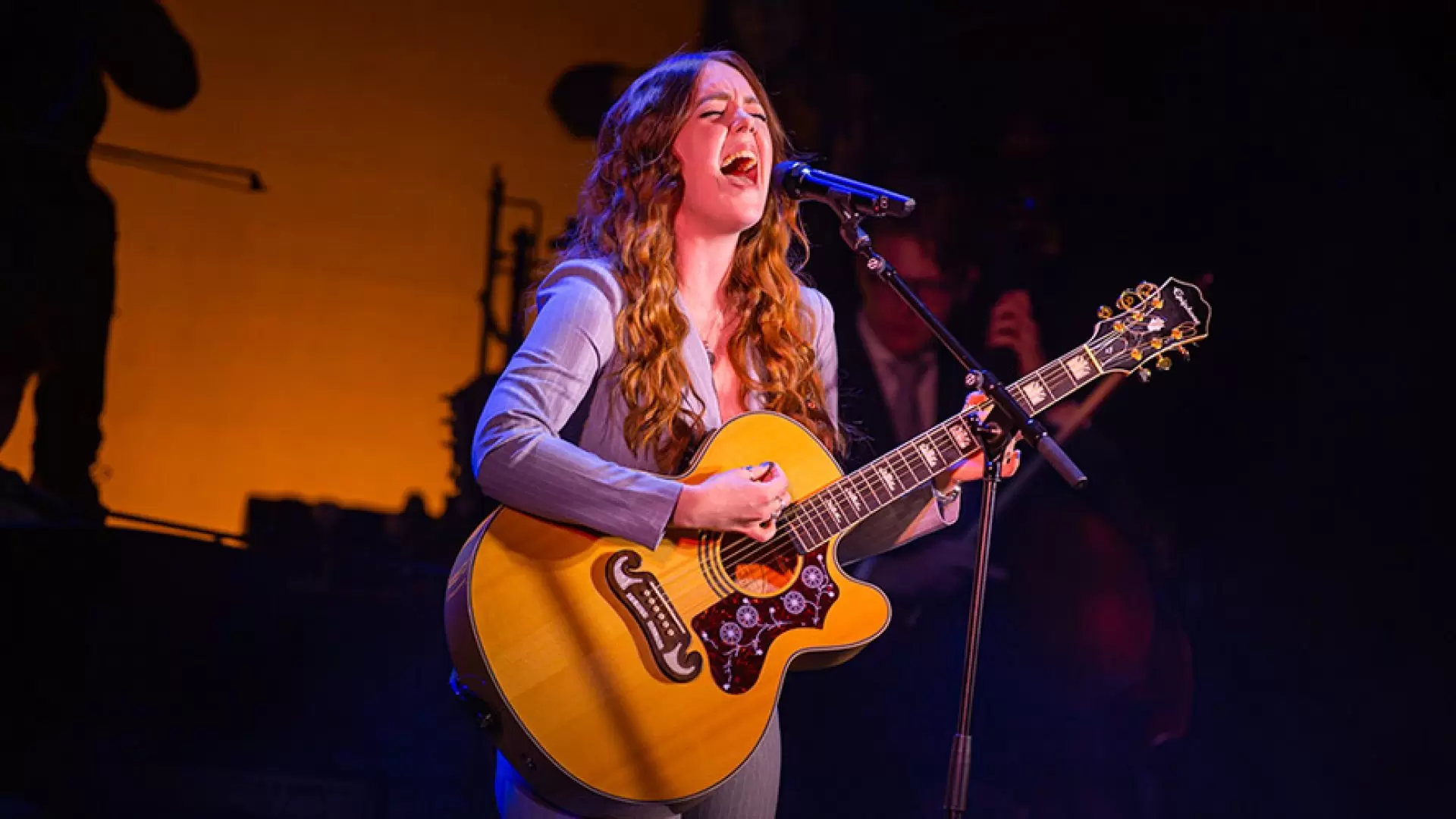 Woman passionately singing and playing acoustic guitar on stage, with warm orange lighting background.