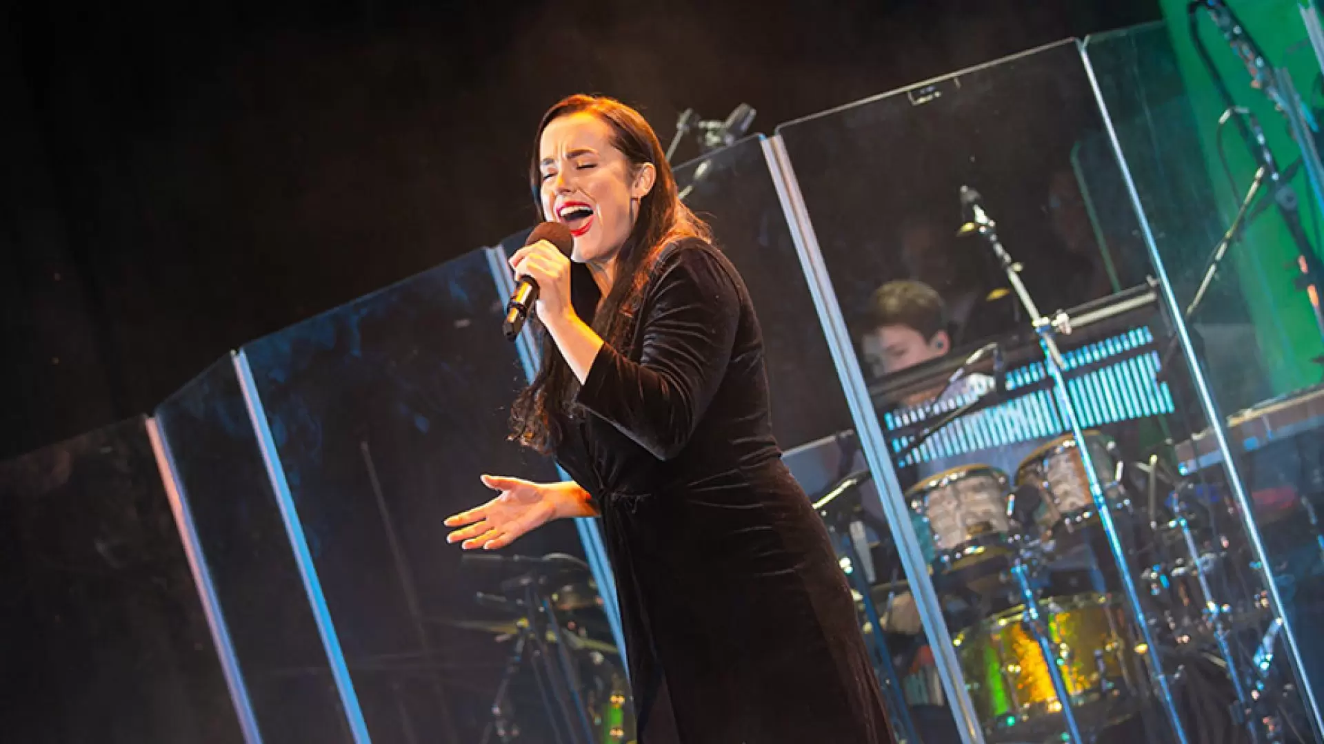 Woman in black dress singing passionately on stage with a microphone, drummer visible behind glass.