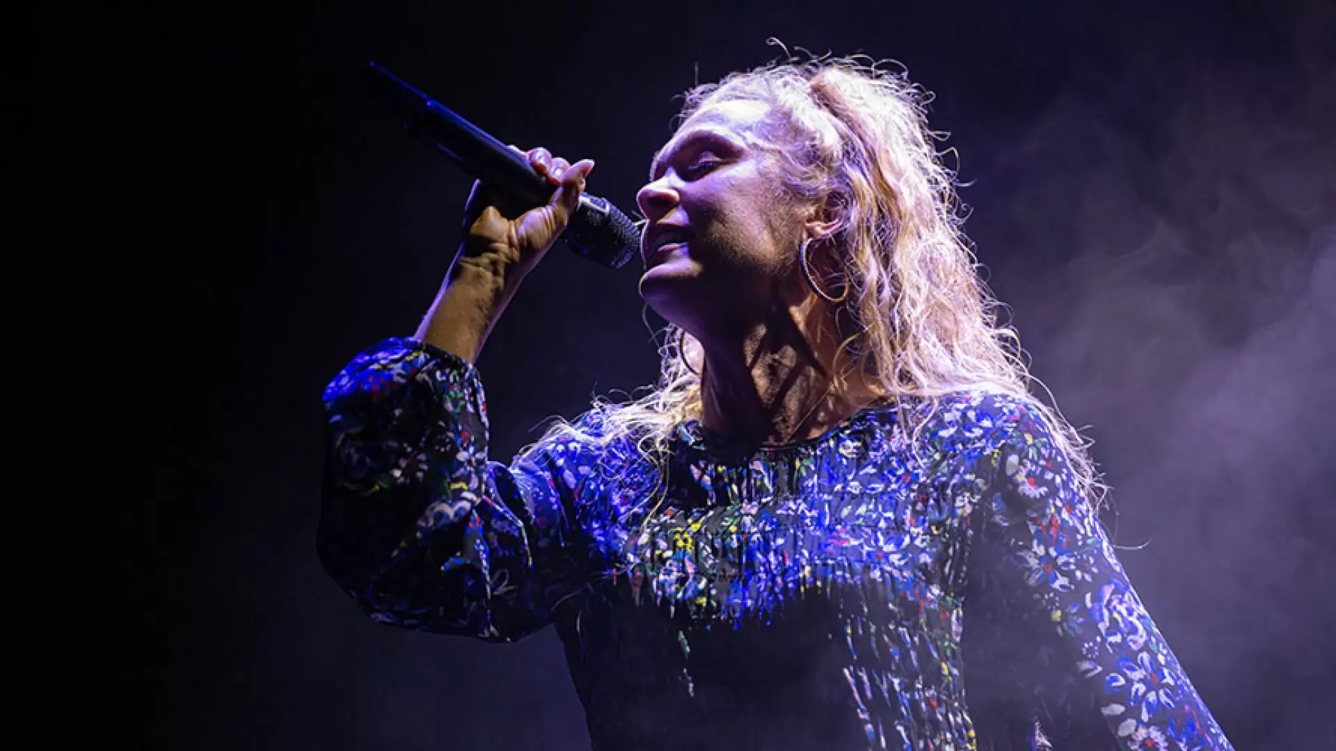 Two actors in theatrical costumes perform on stage with dramatic expressions; a spotlight highlights them.