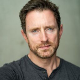 A man with short brown hair and a beard, wearing a grey shirt, against a plain background.