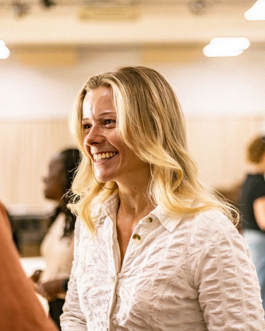 Smiling woman with long blonde hair in a white shirt, standing in a rehearsal room.
