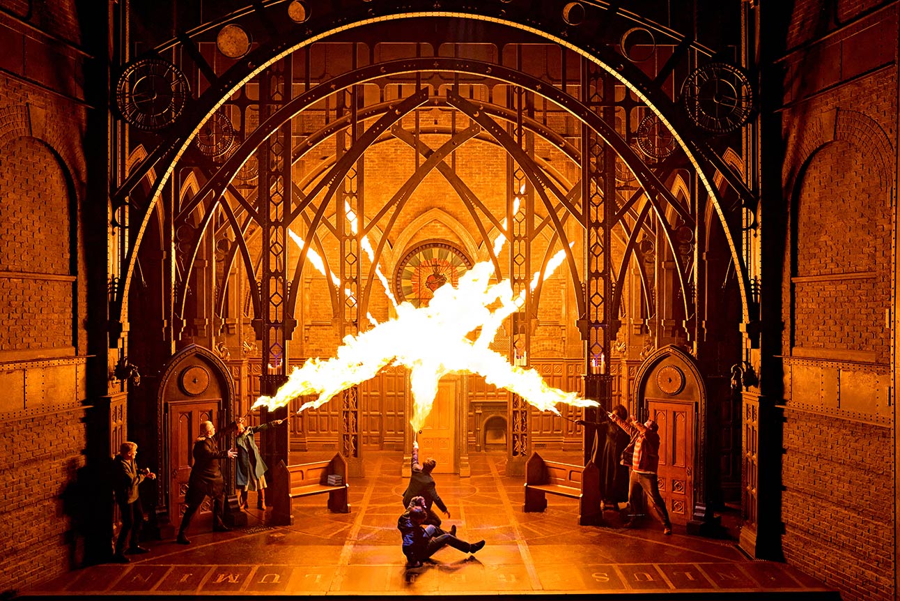 Actors in dramatic battle pose, wearing wizard robes, wands raised, amidst swirling mist and dim lighting.