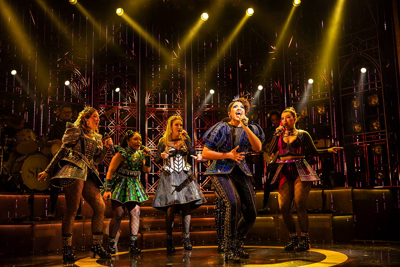 Six performers in colourful Tudor-style costumes sing on stage under bright lights, with a dark backdrop.