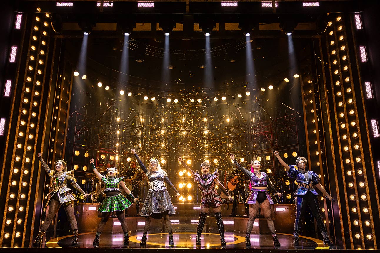 Six performers in colourful, ornate costumes sing and dance energetically on stage against a vibrant backdrop.