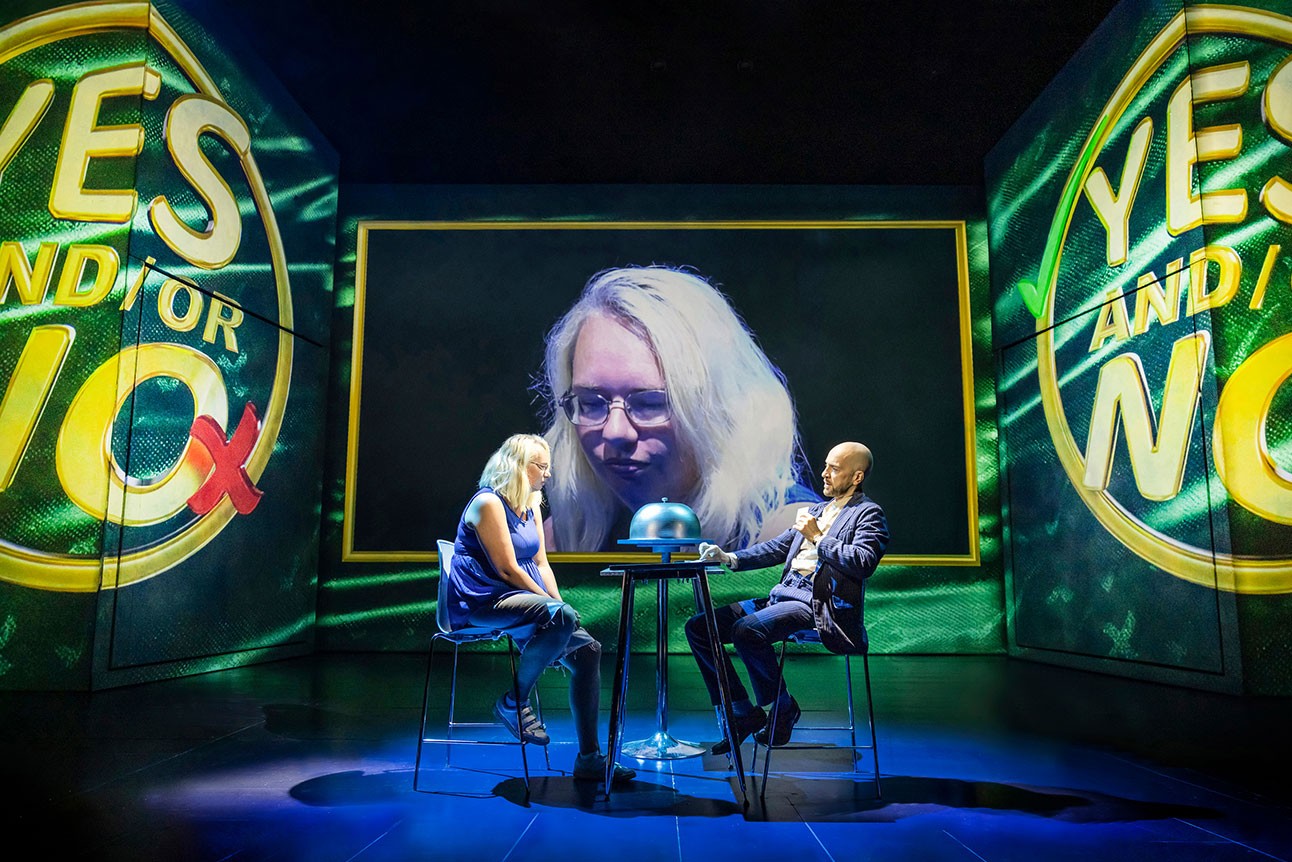 Derren Brown sites a table with a lady. The lady's face is projected onto a large screen behind them.
