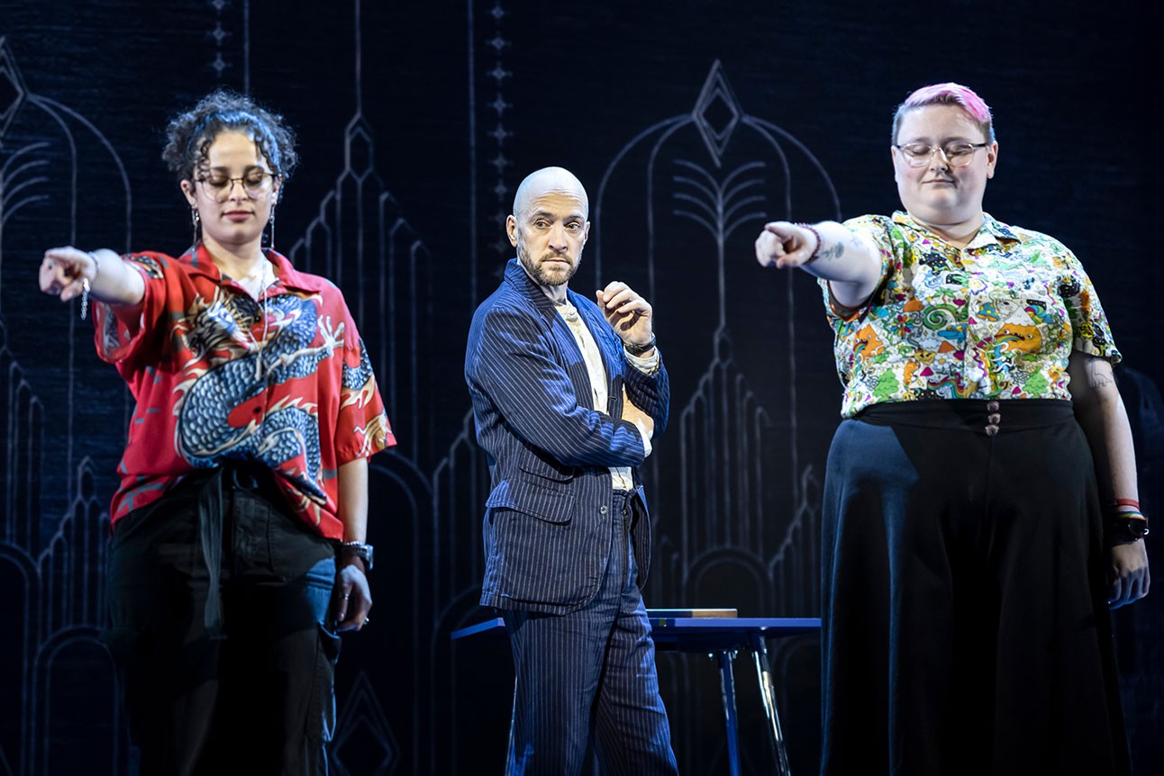Derren Brown stands between two people who are both pointing and both have their eyes closed.