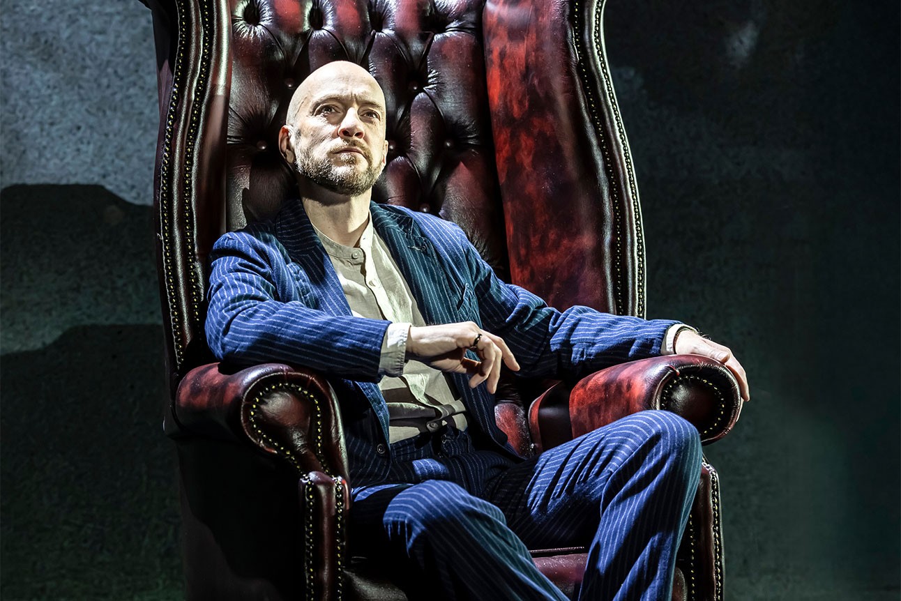 Derren Brown wearing a dark suit sites in a tall armchair