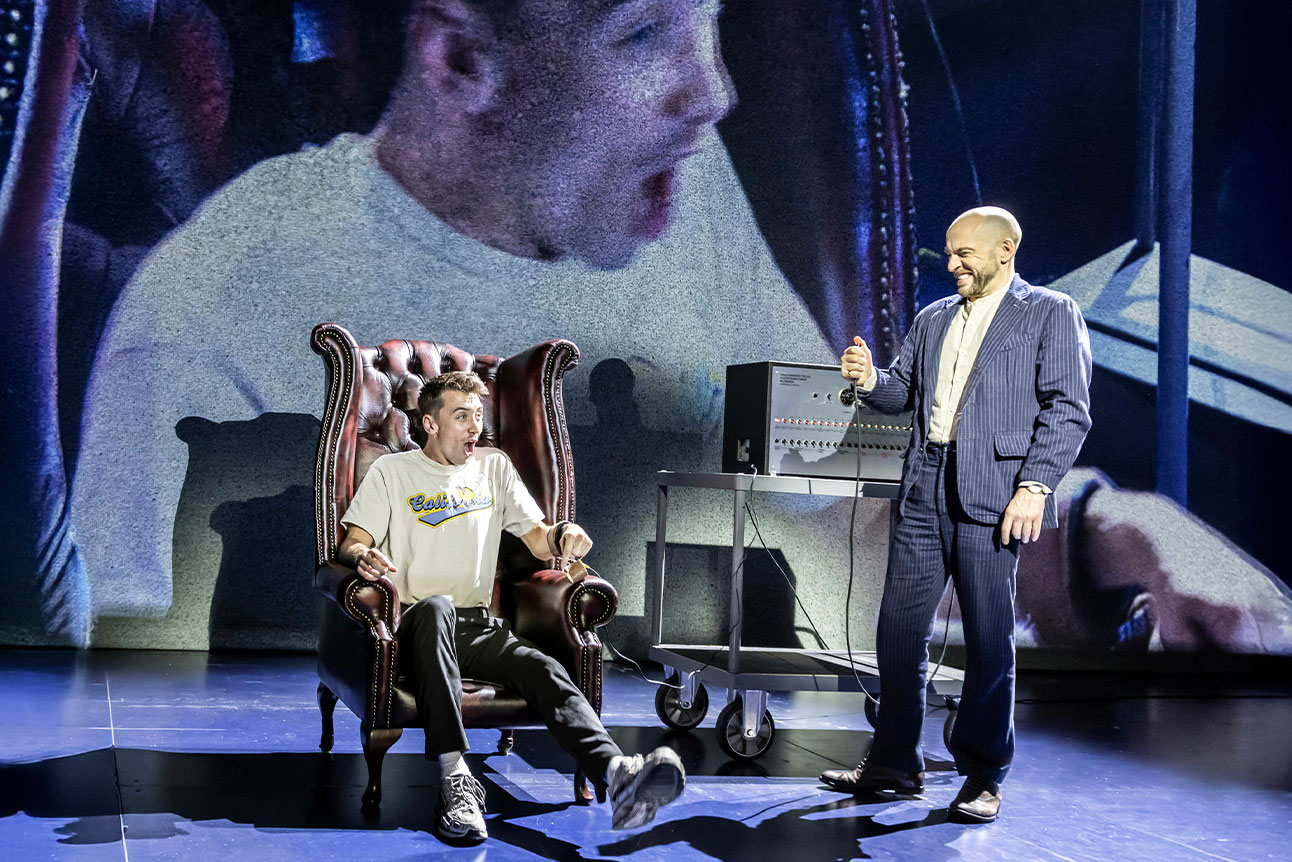 Derren Brown stands on stage, with a audience member sat in a chair