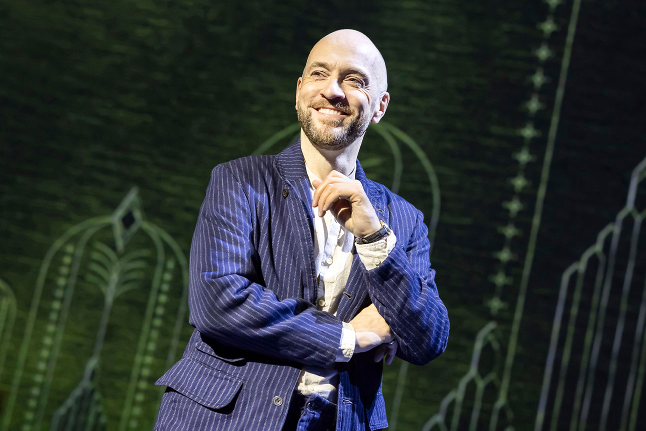 Derren Brown, in a dark suit, smiles on stage against a dark background.