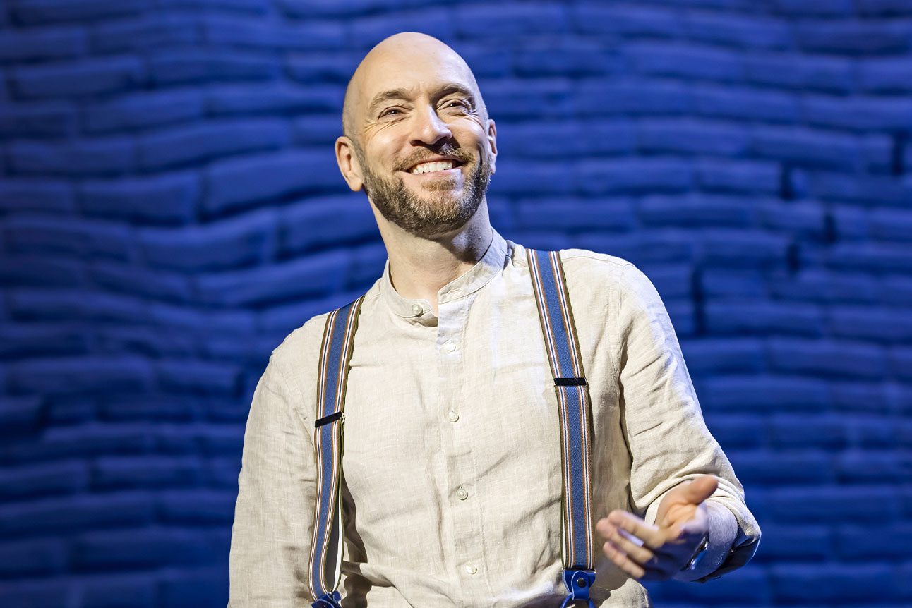 Derren Brown stands on stage gesturing with one hand, wearing a white shirt and braces against a blue backdrop.