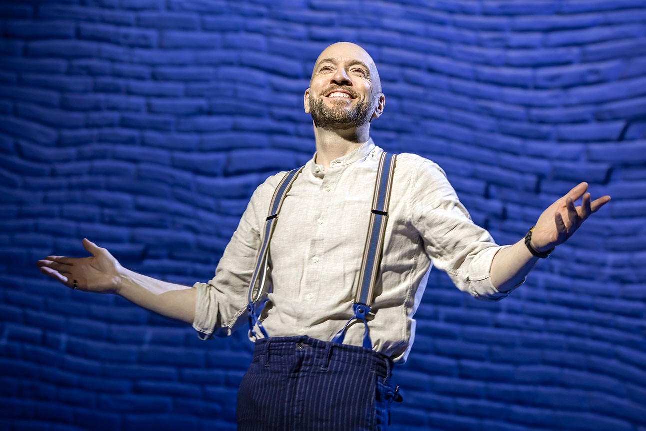 Derren Brown stands on stage, smiling and gesturing with two hands, wearing a white shirt and braces against a blue backdrop.